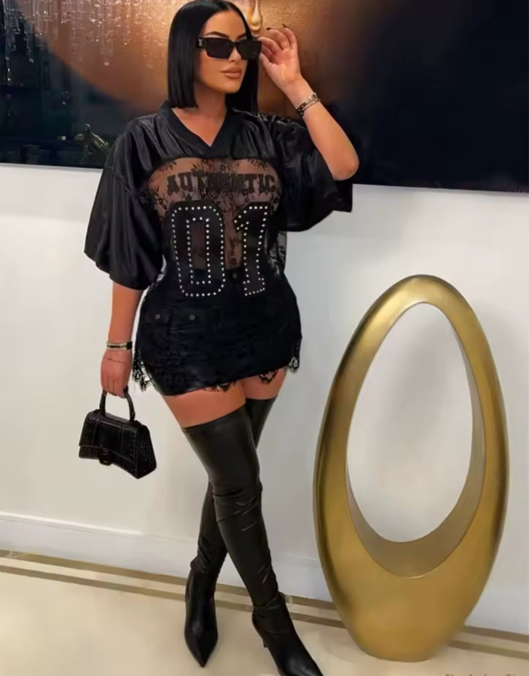 Woman in black outfit with studded details, sunglasses, and high boots standing indoors.