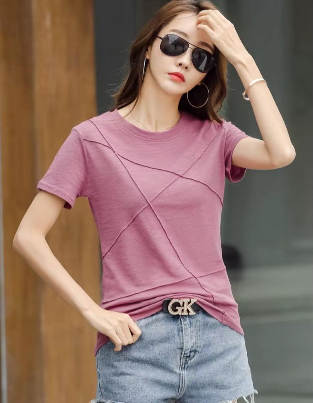 Woman wearing a purple t-shirt and denim shorts with sunglasses, standing against a blurred background.