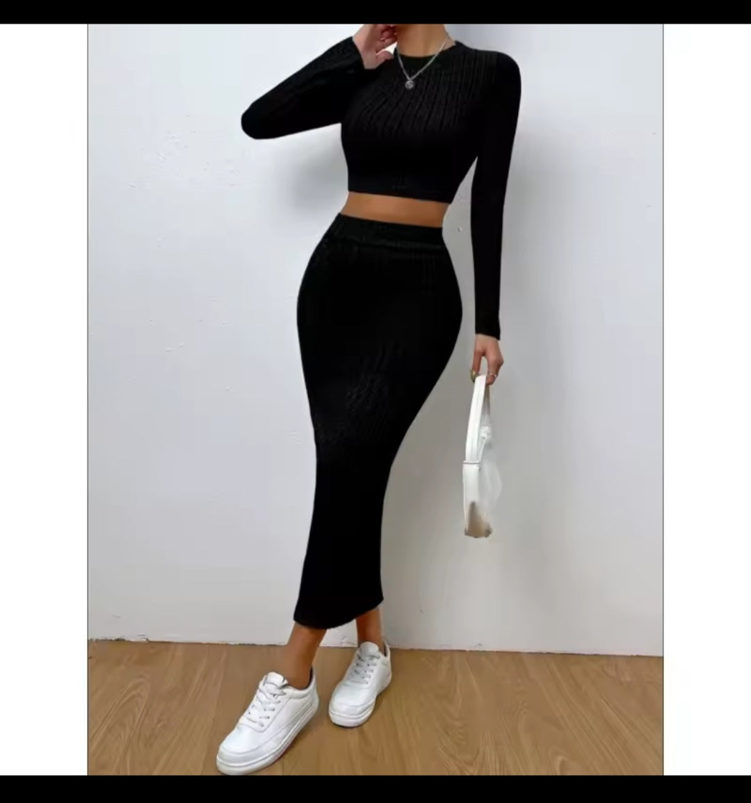 Person wearing a black long-sleeve top and high-waisted pants with white sneakers against a plain background.