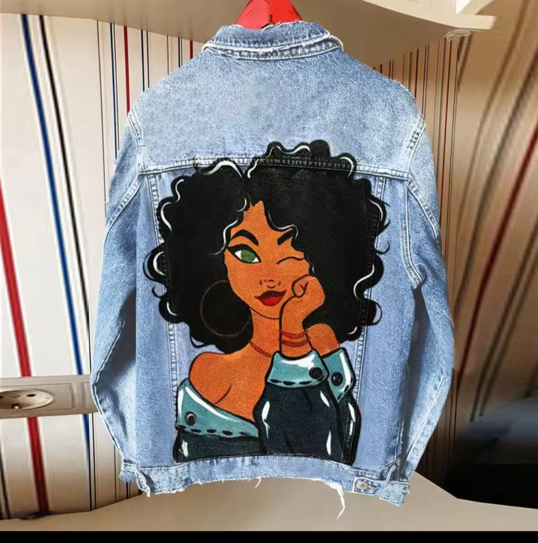 Denim jacket with a colorful graphic of a woman on the back, hanging against a striped wall.