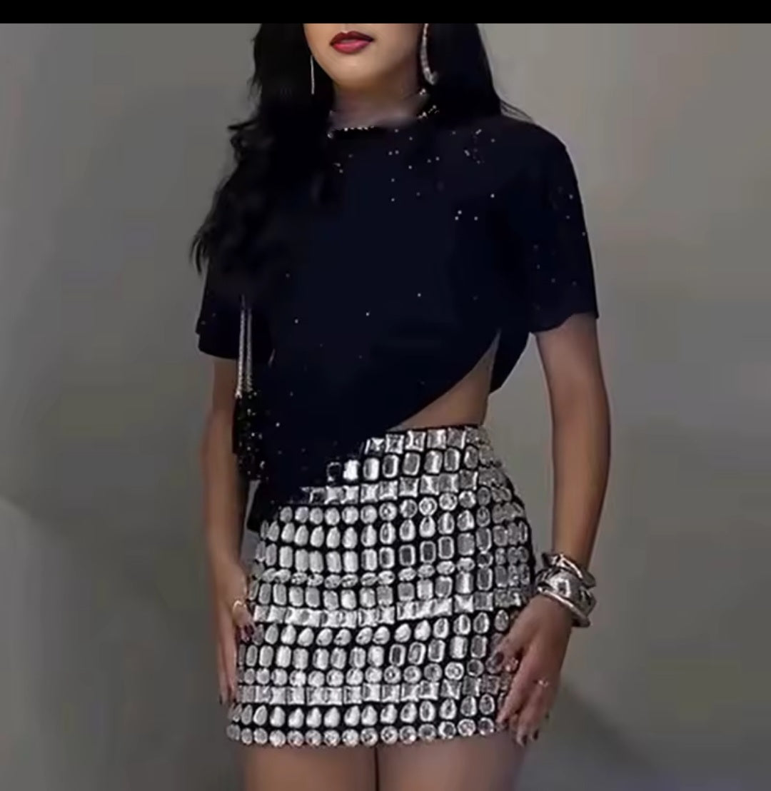 Woman wearing a black top with cut-out design and a silver sequin skirt against a gray background