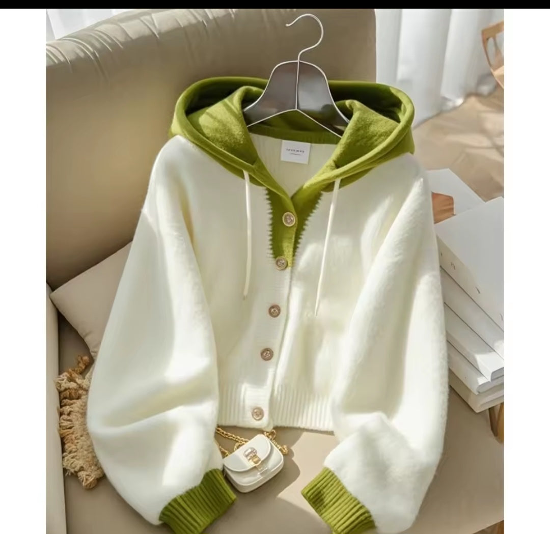 White cardigan with green trim on a hanger against a neutral background