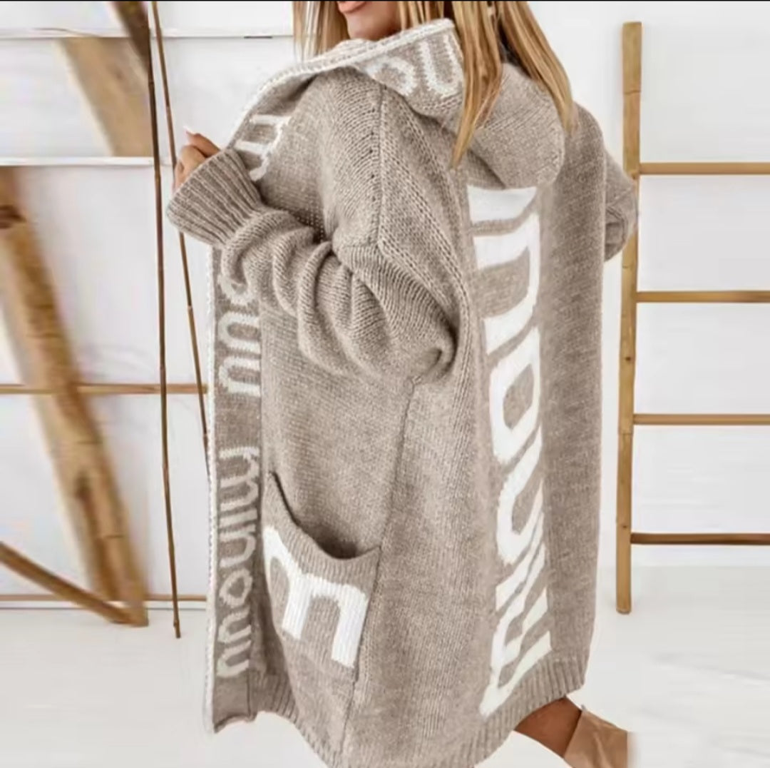 Person wearing a beige oversized sweater with white text, standing against a light-colored wall.